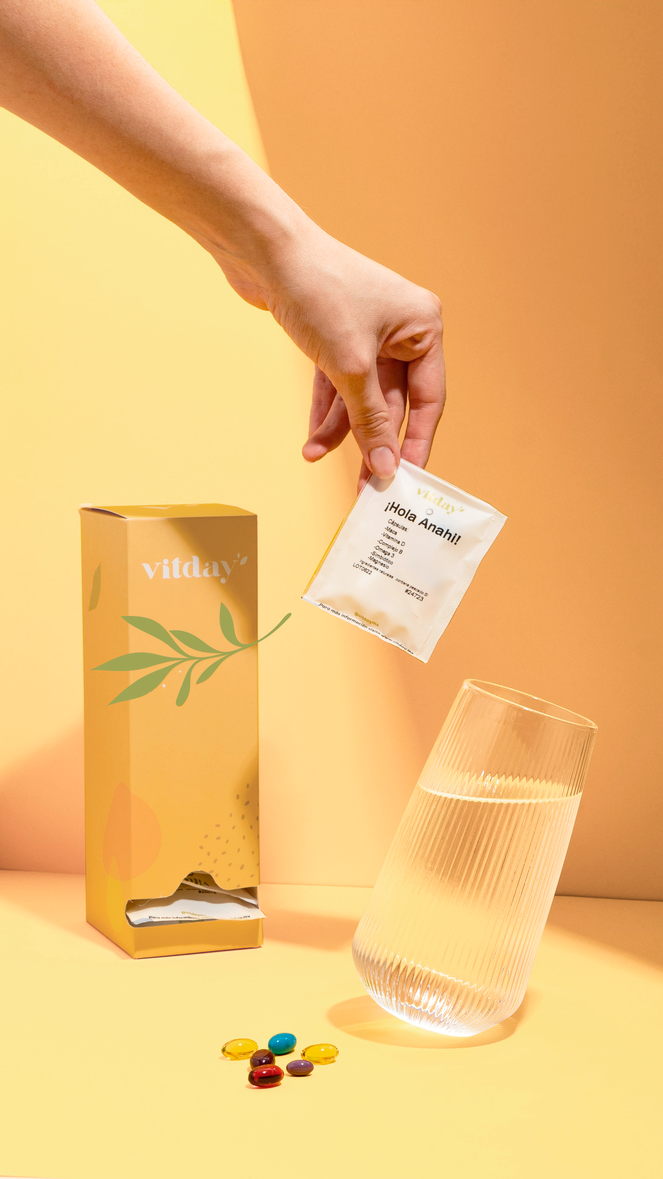 Una mano toma el sobre diario Vitamin junto a su caja y un vaso de agua.